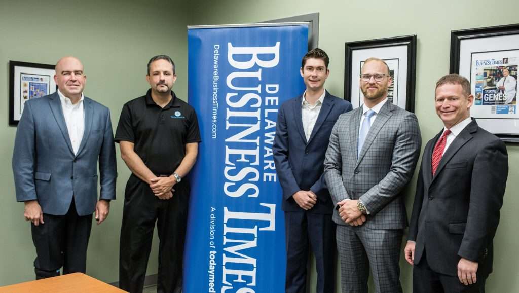 Five men in business attire standing by a Delaware Business Times flyer