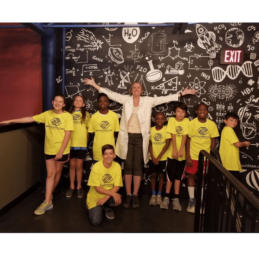 A woman standing in front of a black board with a lot of kids wearing yellow shirts