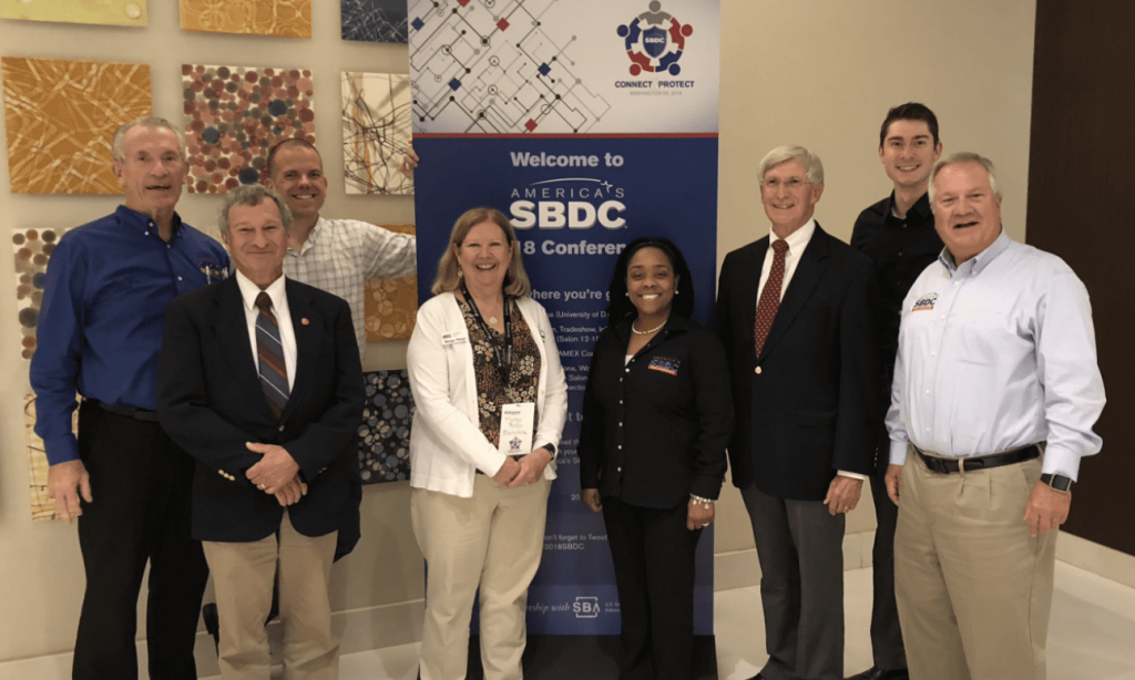 A group of Delaware SBDC staff and advisors standing in front of a poster