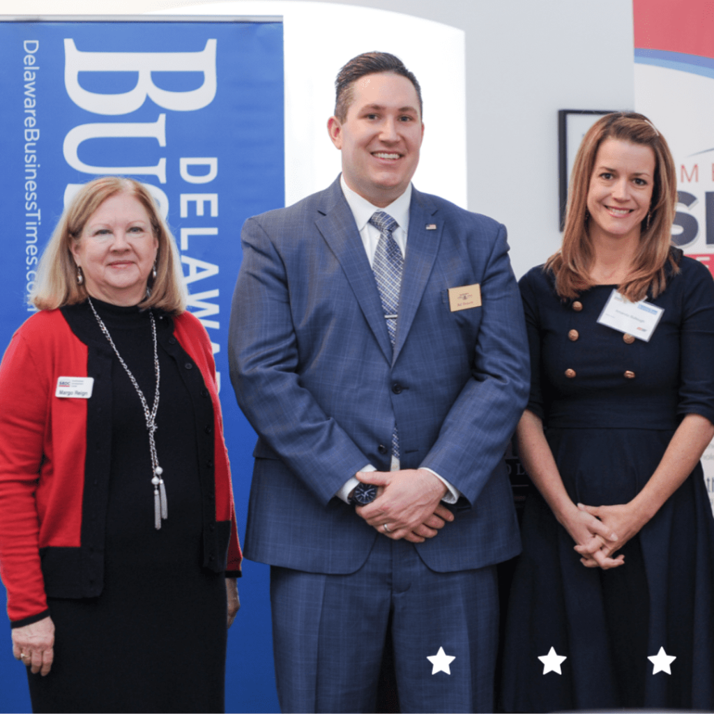 Three people in professional attire standing in front of a Delaware Business Times flier