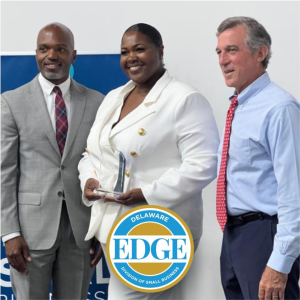 A black woman holding an award amid two men at the Delaware Division of Small Business EDGE grant event.