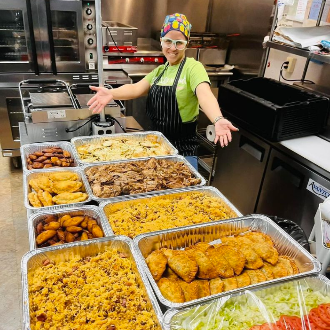 A lady standing in a kitchen with numerous pans of snacks