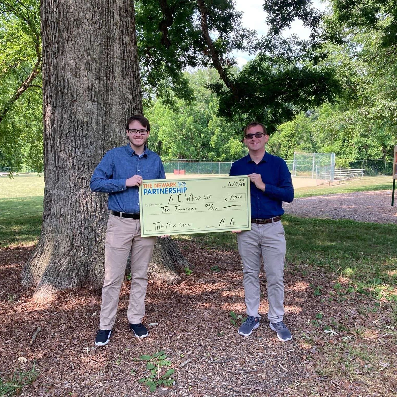 Two men standing in a park holding a large cheque prize